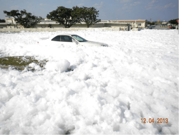Outdoor photo showing a major leak of PFAS foam, with a parked car partially submerged in the foam almost up to its windows.