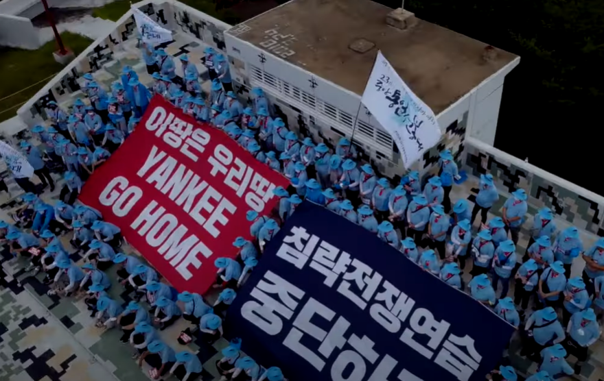 A group of demonstrators at a command post are all dressed in blue uniforms unfurling large banners reading "This land is our land" in Korean, "YANKEE GO HOME" in English, and "Stop practicing for a war of aggression" in Korean.