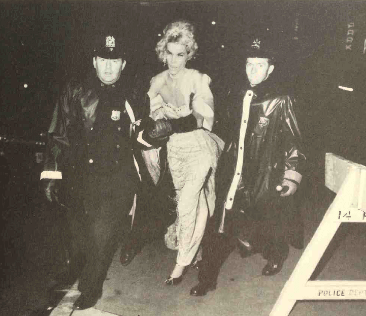 A person in a dress being led by two police looking at the camera