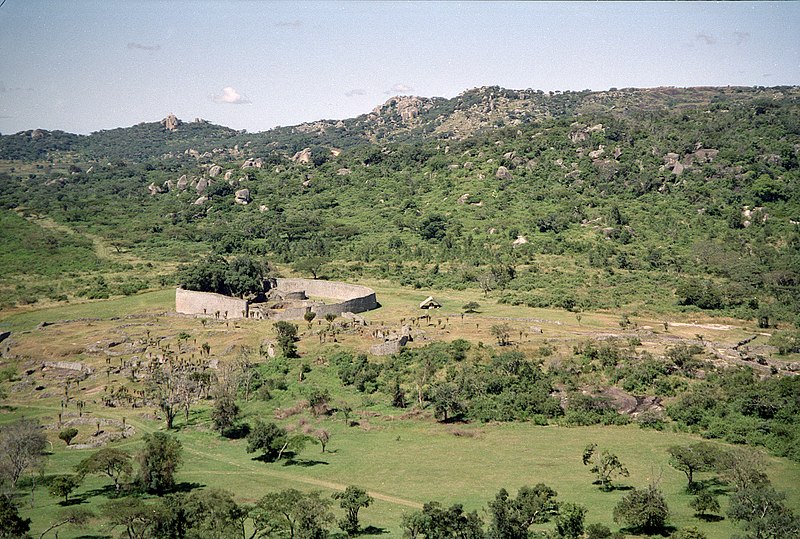 A circular enclosure made of stone walls is situated on a landscape of grass and trees, with hills in the background.