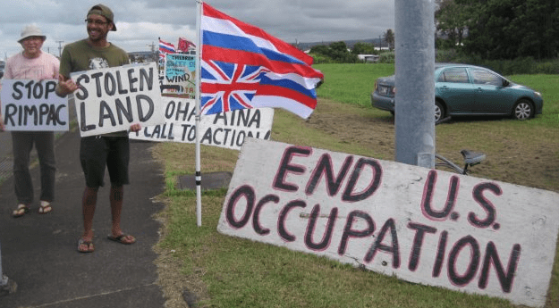 Protestors hold signs reading "STOP RIMPAC", "STOLEN LAND" and "END U.S. OCCUPATION" while standing beside an inverted Hawaiian flag