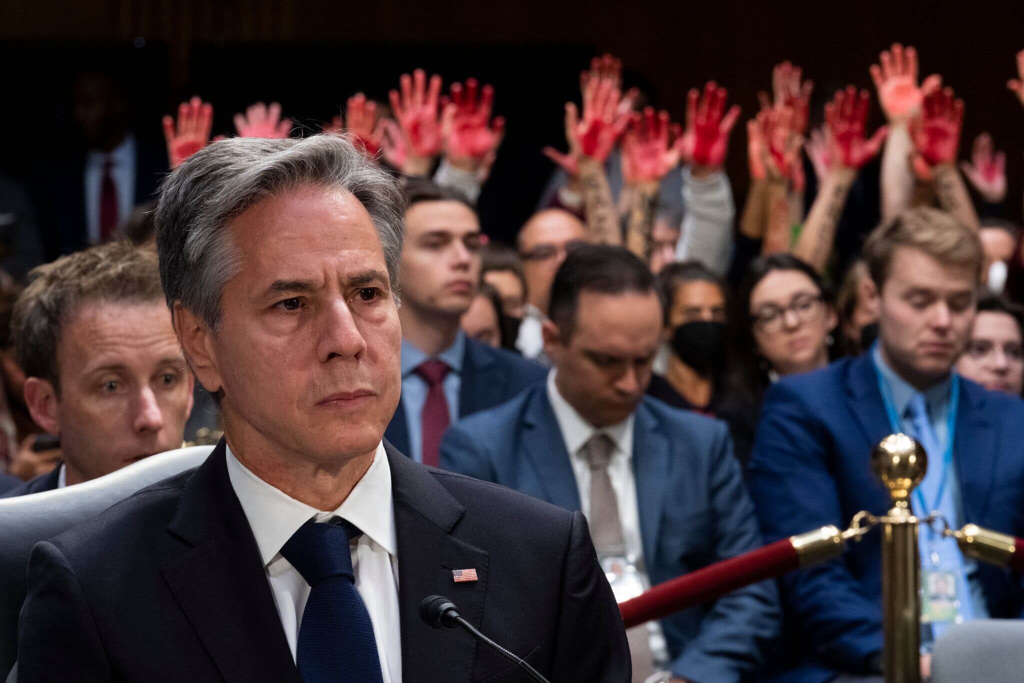 Antony Blinken at a senate hearing on October 31, 2023 regarding a budget request for Israel. Behind him, activists hold up red-painted hands in protest of U.S. support for Israel's bombing campaign in Gaza.