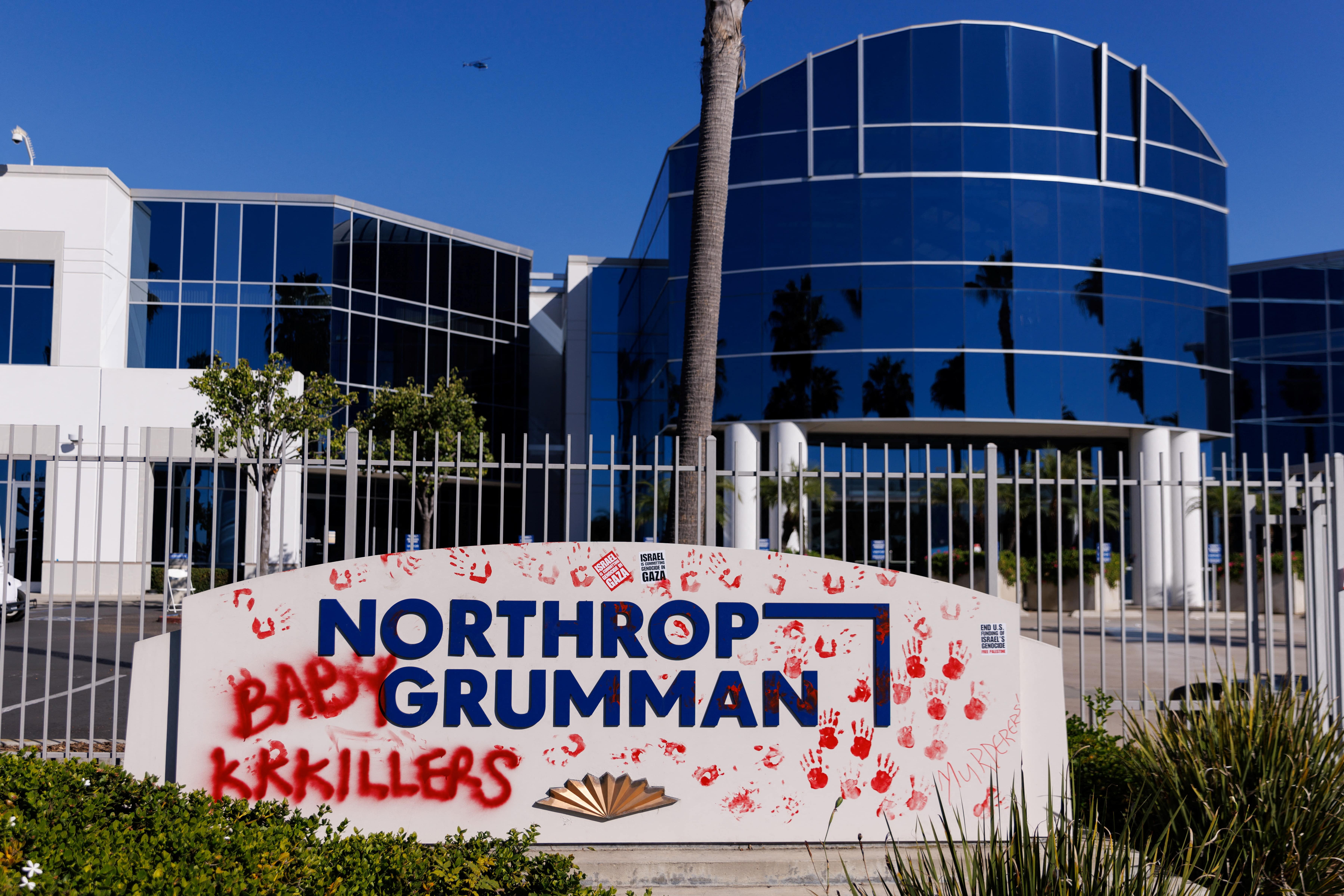 A Northrop Grumman sign covered in red paint depicting red hands and the words "Baby KKKillers" and "Murderers" and stickers reading "Israel is committing genocide in Gaza"