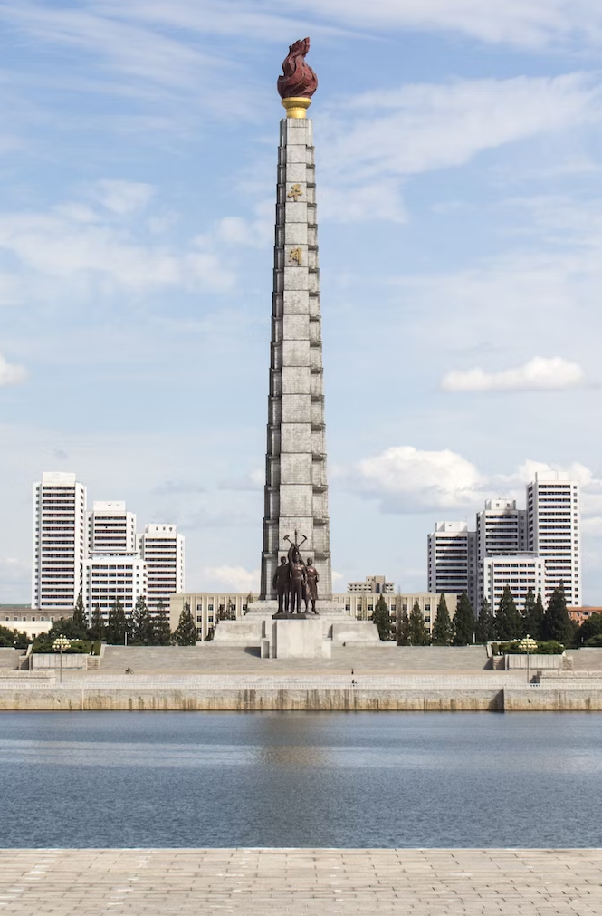 The Tower of the Juche Idea in Pyongyang, DPRK. The word Juche, 주체, is visible on the tower. In front, a statue of a worker, a farmer, and an intellectual hold up a hammer, sickle, and brush, symbol of the Workers' Party of Korea.