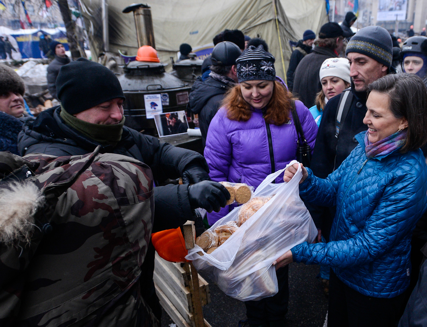 Victoria Nuland smiles as she holds an open bag of snacks for protestors. One person is taking a snack from the bag.