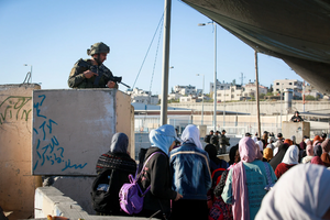 A crowd of Palestinian people walk through a checkpoint where Israeli forces are standing on platforms and pointing guns at the crowd.